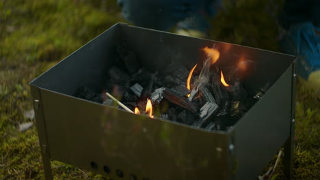 male-camper-is-watching-flames-in-chargrill-colas-are-burning-preparing-for-cooking-meat-resting-in-tent-camp-in-forest